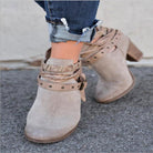 Beige suede ankle boots with braided straps and studded details, paired with rolled-up denim jeans. Fashionable women's footwear on pavement.