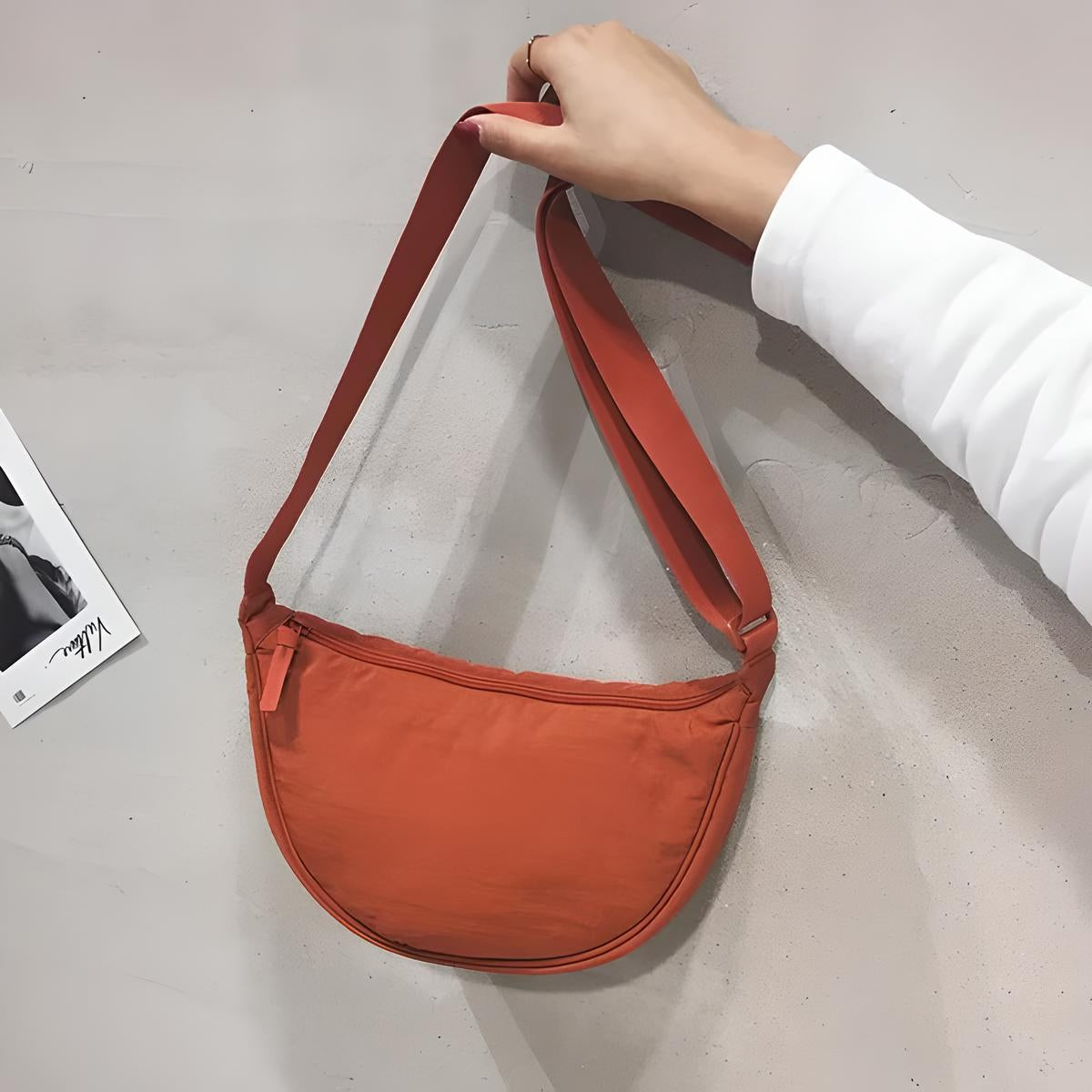 Orange crescent-shaped crossbody bag with adjustable strap, held by a hand against a neutral background. Stylish, minimalist fashion accessory.