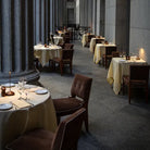 Elegant outdoor restaurant with candlelit tables, brown chairs, and stone columns, creating a romantic dining atmosphere. Fine dining, al fresco, evening.