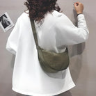 Woman in white sweatshirt with curly hair, wearing an olive green crossbody bag, standing against a gray wall. Fashionable casual outfit.