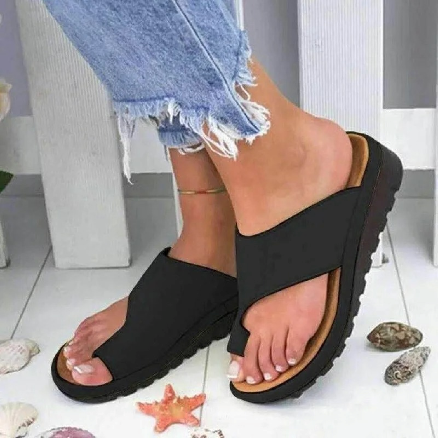 Black slip-on sandals with thick soles on feet, paired with frayed hem jeans. Beach-themed decor with seashells and starfish on white floor.