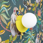 Modern wall lamp with a round white globe and yellow base, set against a vibrant jungle-themed wallpaper featuring monkeys and tropical birds.