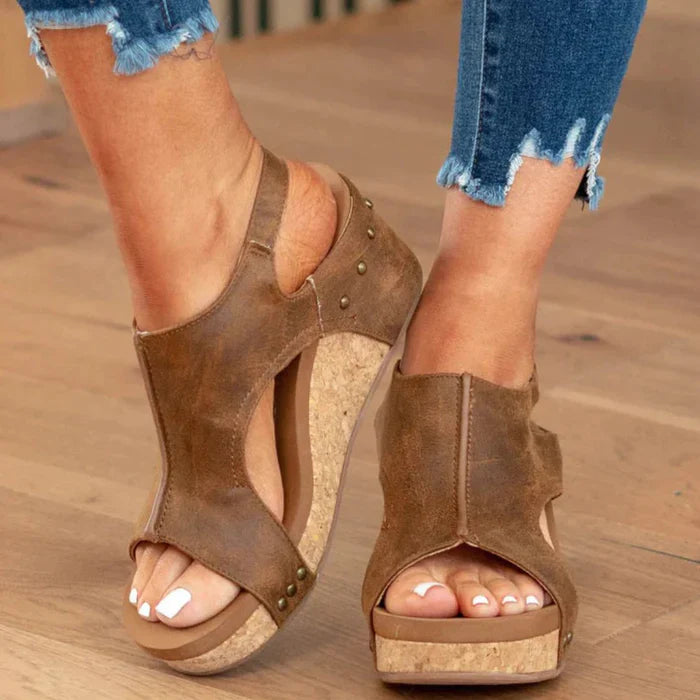 Women's brown leather wedge sandals with open toe design, cork platform, and stud accents, paired with distressed denim jeans on wooden floor.