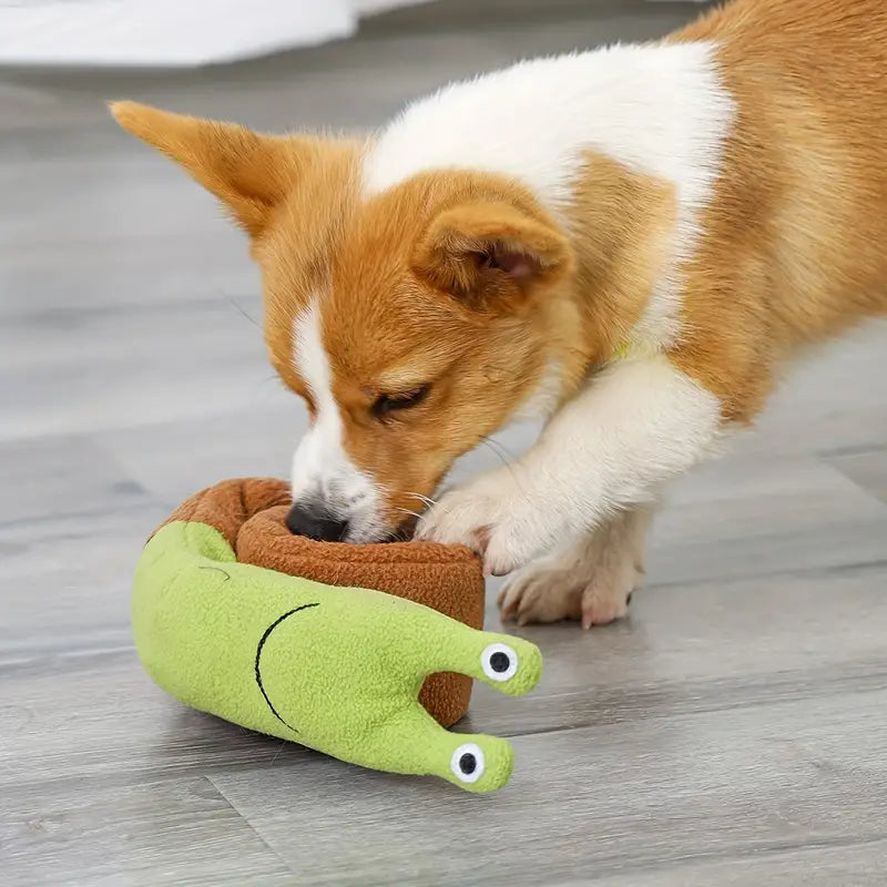 Corgi puppy playing with a green snail-shaped plush toy on a wooden floor. Cute dog, pet toy, playful puppy, indoor pet activity.