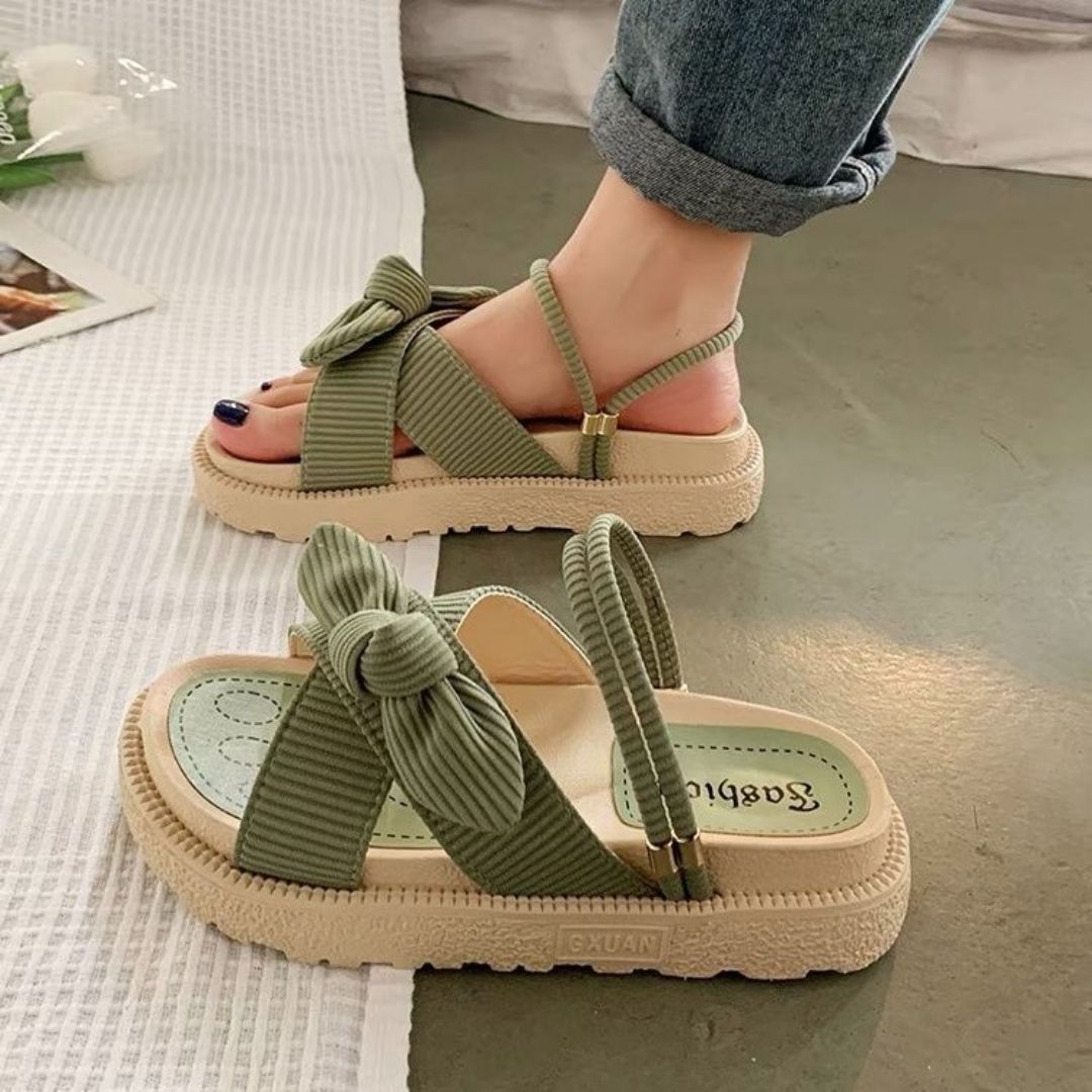 Green bow strap sandals with cushioned sole, worn by a person in jeans. Stylish summer footwear, perfect for casual outings and comfort.