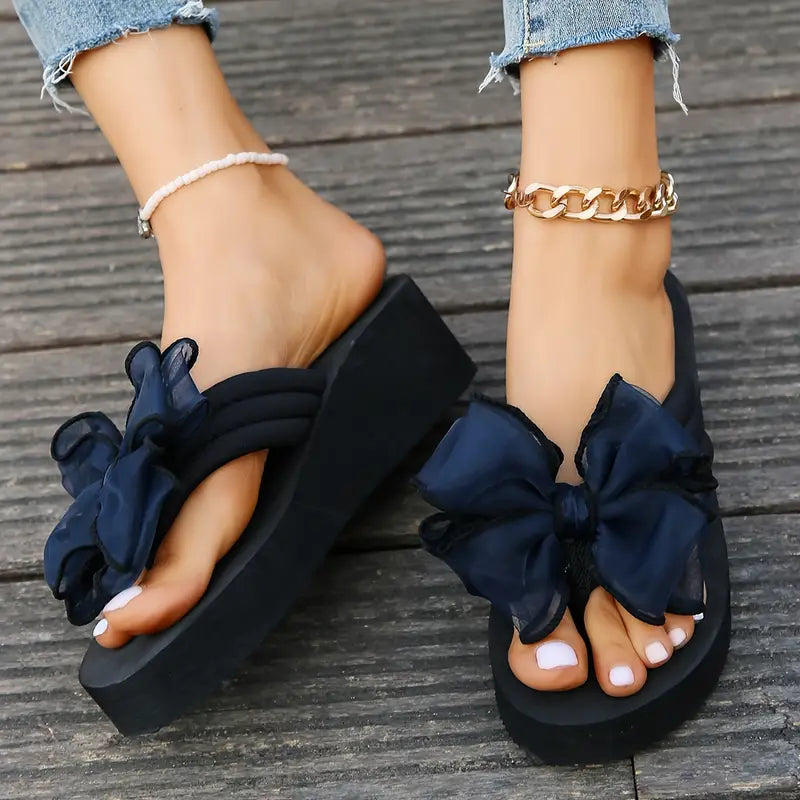 Women's black platform sandals with large navy bows, worn with anklets and jeans. Fashionable summer footwear, stylish casual look.