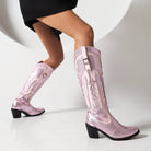 Pink cowboy boots with intricate embroidery, mid-heel design, and pointed toes, worn by a model in a black dress. Fashion footwear, Western style.