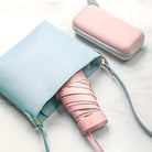 Pastel blue handbag with pink strap, featuring a compact pink umbrella and matching pink wallet on a marble surface. Fashion accessories, stylish design.