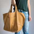 Woman holding large tan leather tote bag with front pocket, wearing casual green top and blue jeans. Fashionable, spacious handbag for everyday use.