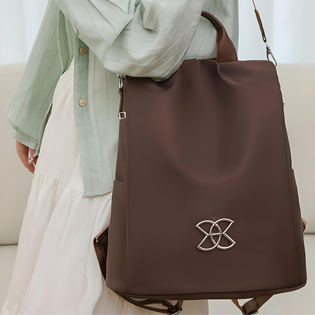 Elegant brown leather backpack with silver logo, worn by a person in a light green shirt and white skirt. Stylish fashion accessory for women.