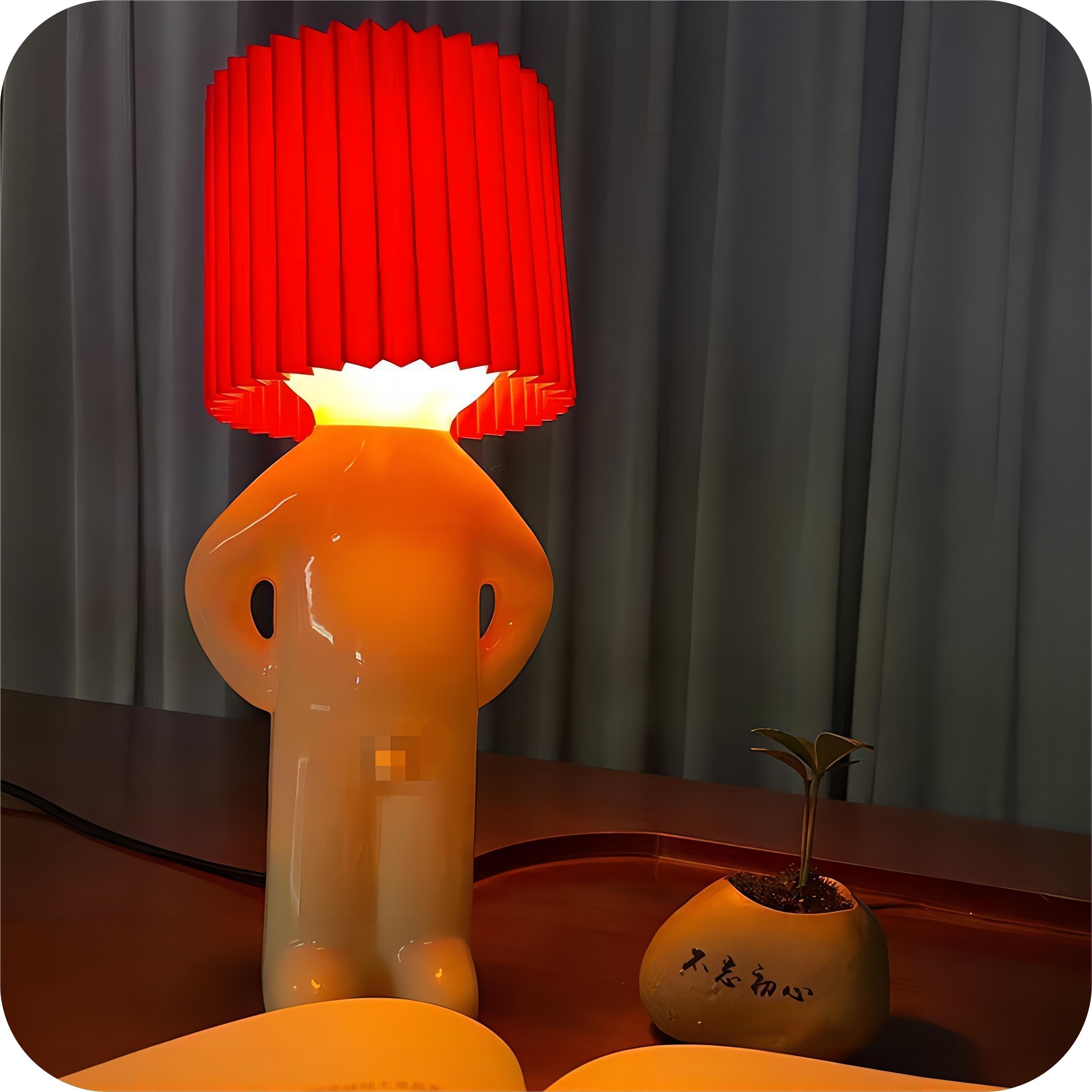 Quirky table lamp with a red pleated lampshade shaped like a person, next to a small potted plant on a wooden desk, modern home decor.