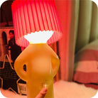 Quirky yellow lamp with a red pleated lampshade, shaped like a person, illuminating a cozy bedroom setting. Unique home decor lighting accessory.