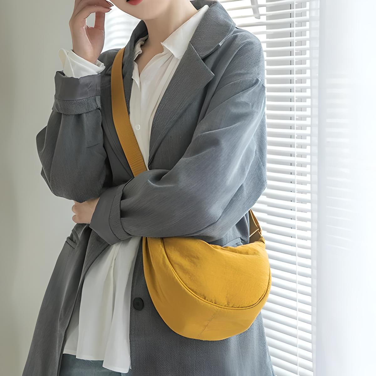 Woman in gray blazer holding a mustard yellow crossbody bag, standing by window blinds. Fashionable casual outfit, stylish accessory, modern look.