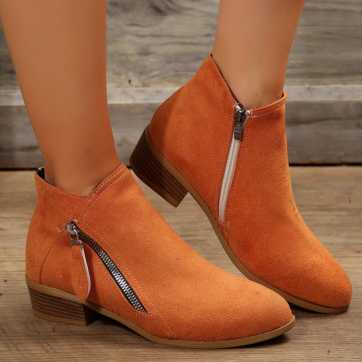 Orange suede ankle boots with side zippers, low block heels, and wooden flooring background. Stylish women's footwear, perfect for fall fashion.