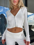 Woman in stylish white ribbed knit zip-up sweater and matching pants, standing outdoors with ocean view. Fashionable casual outfit, beachwear.