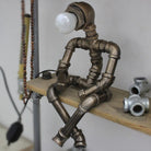 Industrial pipe lamp sculpture with bulb head, sitting on a wooden shelf. Unique steampunk decor, metal pipe art, creative home lighting.