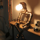 Industrial pipe lamp shaped like a sitting human figure, featuring a glowing bulb head. Unique steampunk decor, perfect for vintage home lighting.