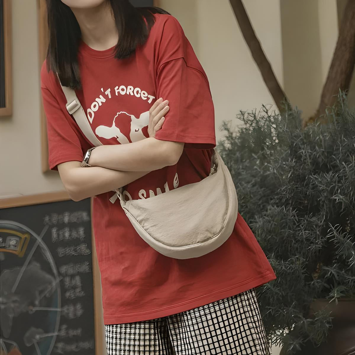 Woman in red graphic t-shirt with "Don't Forget" text, wearing a beige crossbody bag, standing in front of a chalkboard and greenery. Fashion, casual style.