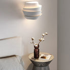 Modern white wall sconce illuminating a minimalist bedroom with a round side table, brown vase, and cotton plant; contemporary home decor lighting.