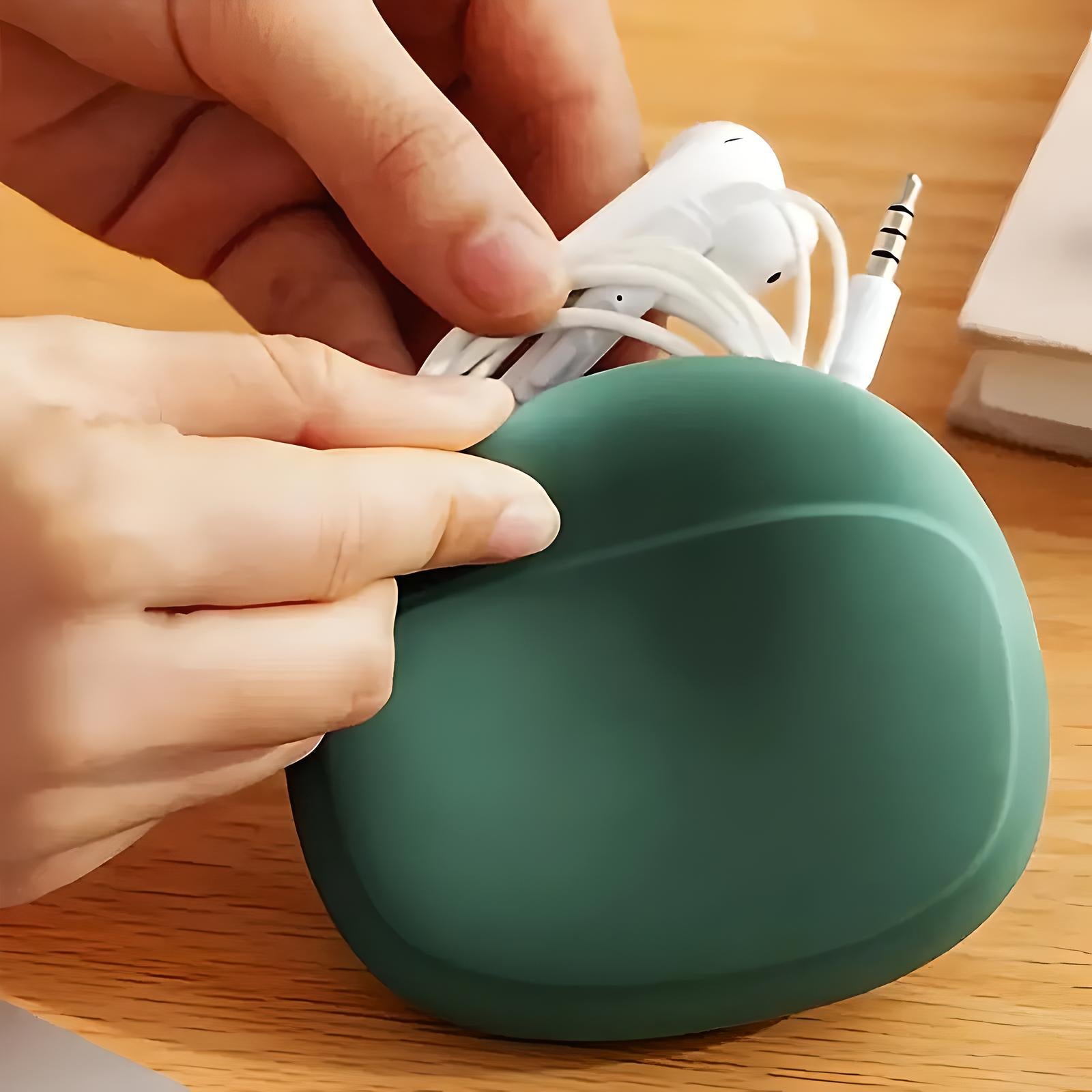 Person holding a green silicone earphone case with white earphones, on a wooden table. Compact earbud organizer, cable management accessory.