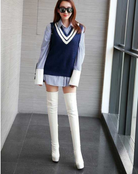 Fashionable woman in blue sweater vest over striped shirt, wearing white thigh-high boots and sunglasses, standing indoors. Trendy women's outfit.