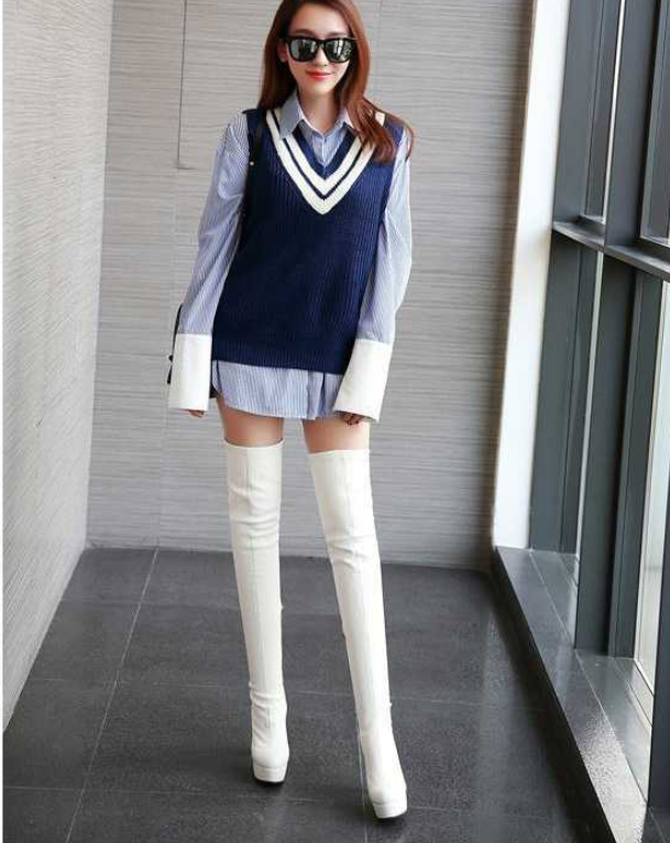 Fashionable woman in blue sweater vest over striped shirt, wearing white thigh-high boots and sunglasses, standing indoors. Trendy women's outfit.