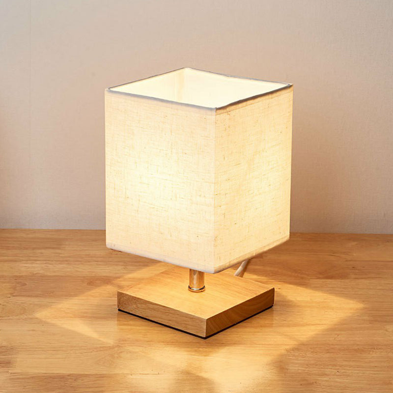 Square table lamp with beige fabric shade on wooden base, emitting warm light. Ideal for modern home decor, bedroom, or living room ambiance.