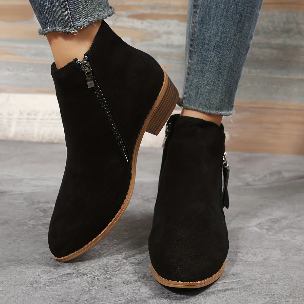 Black suede ankle boots with side zippers, worn with distressed denim jeans. Stylish women's footwear, perfect for casual fall fashion.