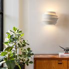 Modern white wall sconce with layered design illuminating a minimalist room, featuring a wooden cabinet and lush green indoor plant.