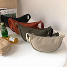 Four stylish crossbody bags in beige, olive, and rust colors on a white table, next to a Perrier bottle and magazines. Fashionable accessory display.