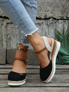 Women's black espadrille wedge sandals with brown ankle straps, paired with distressed skinny jeans. Stylish summer footwear on wooden floor.