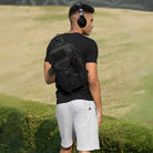 Man wearing black tactical backpack and headphones, standing outdoors on grass. Casual summer outfit with black t-shirt and white shorts.