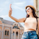 Woman using portable handheld fan outdoors; wearing sunglasses and hat, smiling in sunny weather. Summer cooling device, travel accessory.