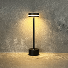 Modern black LED table lamp with circular base and cylindrical shade, illuminating a textured concrete wall. Ideal for minimalist home decor lighting.