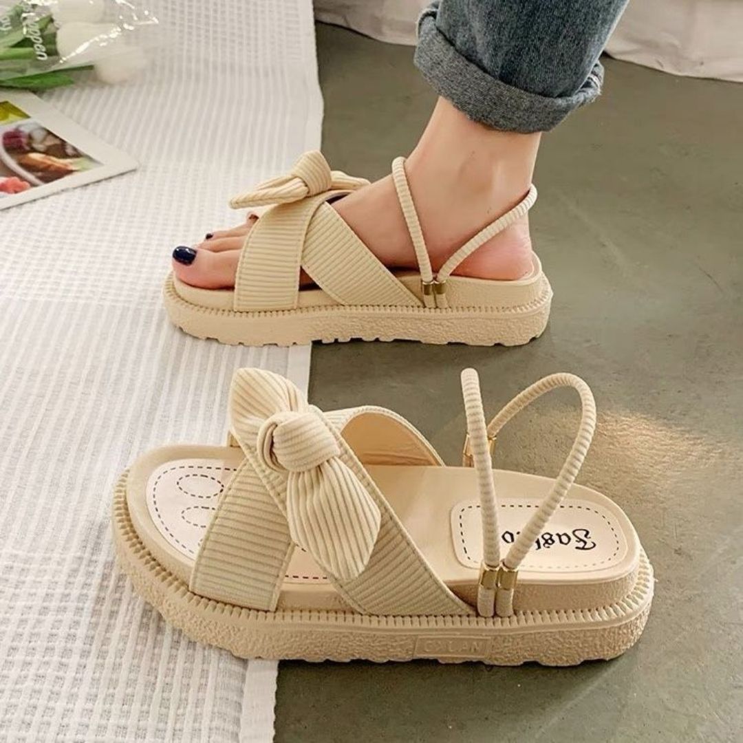 Beige women's sandals with bow detail, flat sole, and ankle strap, worn with rolled-up jeans. Stylish summer footwear on a textured surface.