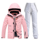 Pink women's ski jacket with hood and white snow pants set, waterproof winter sportswear.