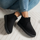 Black suede ankle boots with faux fur lining, worn with blue jeans. Stylish winter footwear, slip-on design, perfect for casual outfits.