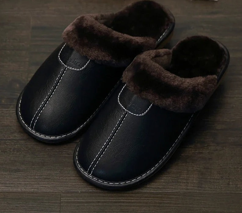 Black leather slippers with plush brown lining on dark wood floor; cozy indoor footwear, perfect for winter warmth and comfort.