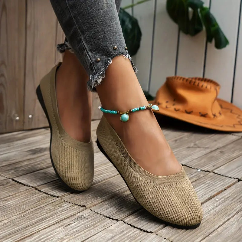 Beige women's slip-on flats with textured fabric, paired with a turquoise anklet. Casual footwear on wooden floor, denim jeans, and a cowboy hat nearby.