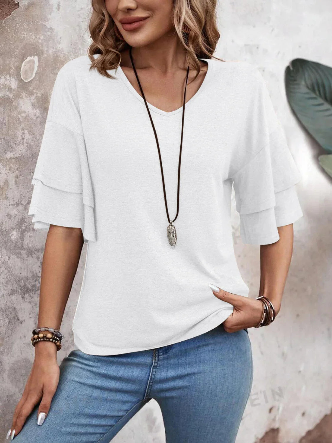 Woman wearing a white ruffle sleeve blouse with a long necklace and blue jeans, standing against a textured wall. Fashionable casual outfit.