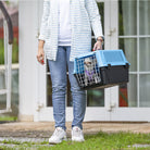 Person carrying a blue and black pet carrier with a small dog inside, standing outside a house. Pet travel, dog crate, outdoor setting.