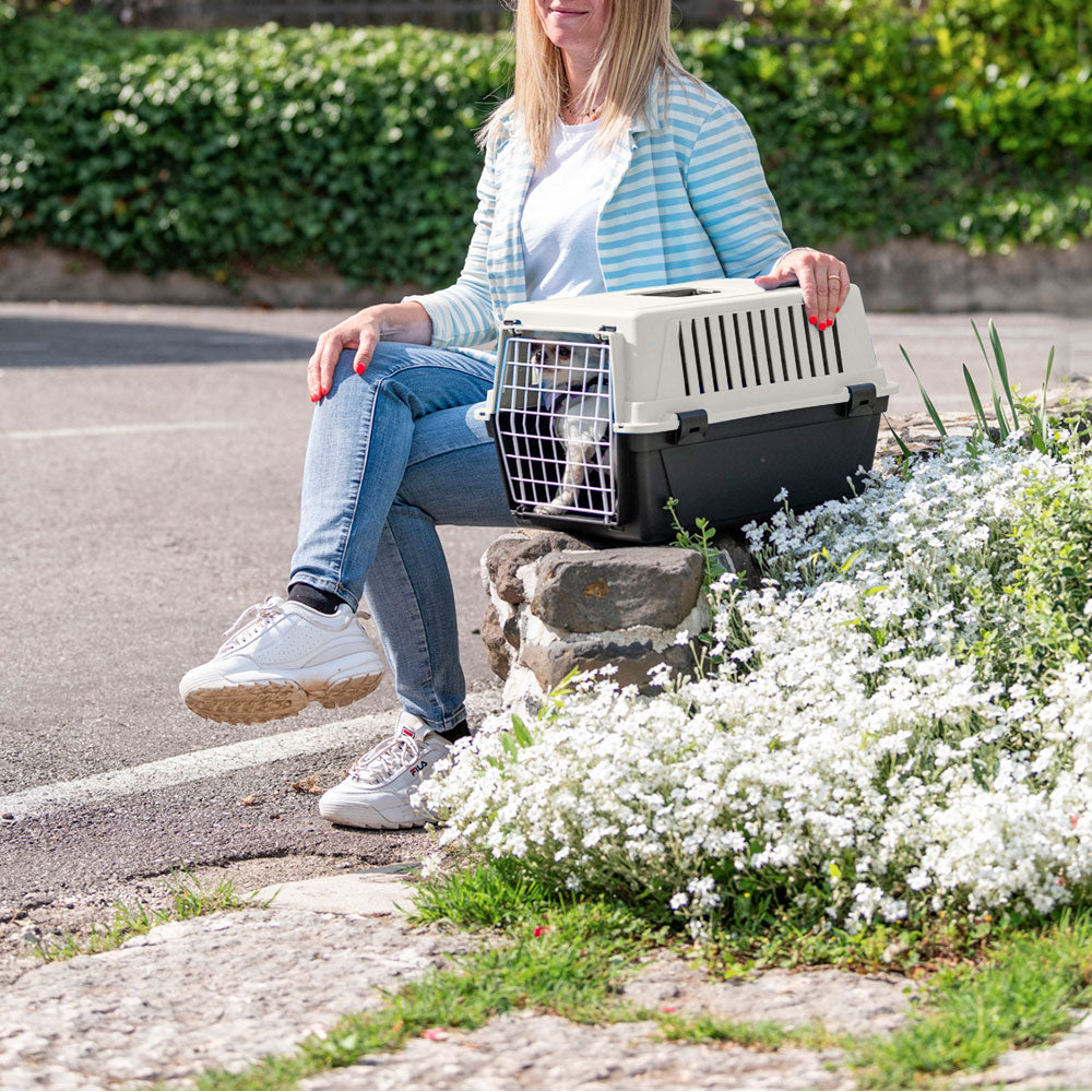 Woman sitting on a stone bench with a pet carrier, surrounded by white flowers, wearing casual jeans and sneakers. Outdoor pet travel concept.