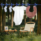 Baby onesie, socks, and bib hanging on a clothesline in a forest setting with a wooden chair and table nearby. Outdoor laundry drying scene.