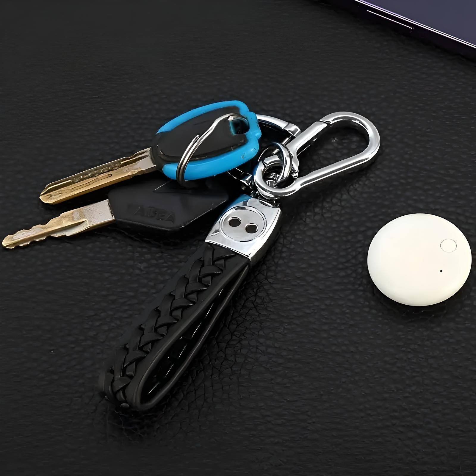 Keychain with braided leather strap, two keys, and a blue key fob on a black surface. Nearby is a small white Bluetooth tracker.