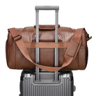 Brown leather duffel bag with shoulder strap on a silver hard-shell suitcase, travel luggage, stylish carry-on, durable travel gear.