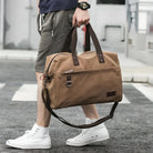 Man carrying stylish brown canvas duffel bag with leather straps, wearing casual shorts and white sneakers. Perfect for travel and gym use.