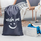 Large black laundry bag with "wash me" text and smiley face, placed in a modern bedroom setting. Ideal for organizing clothes, home decor.