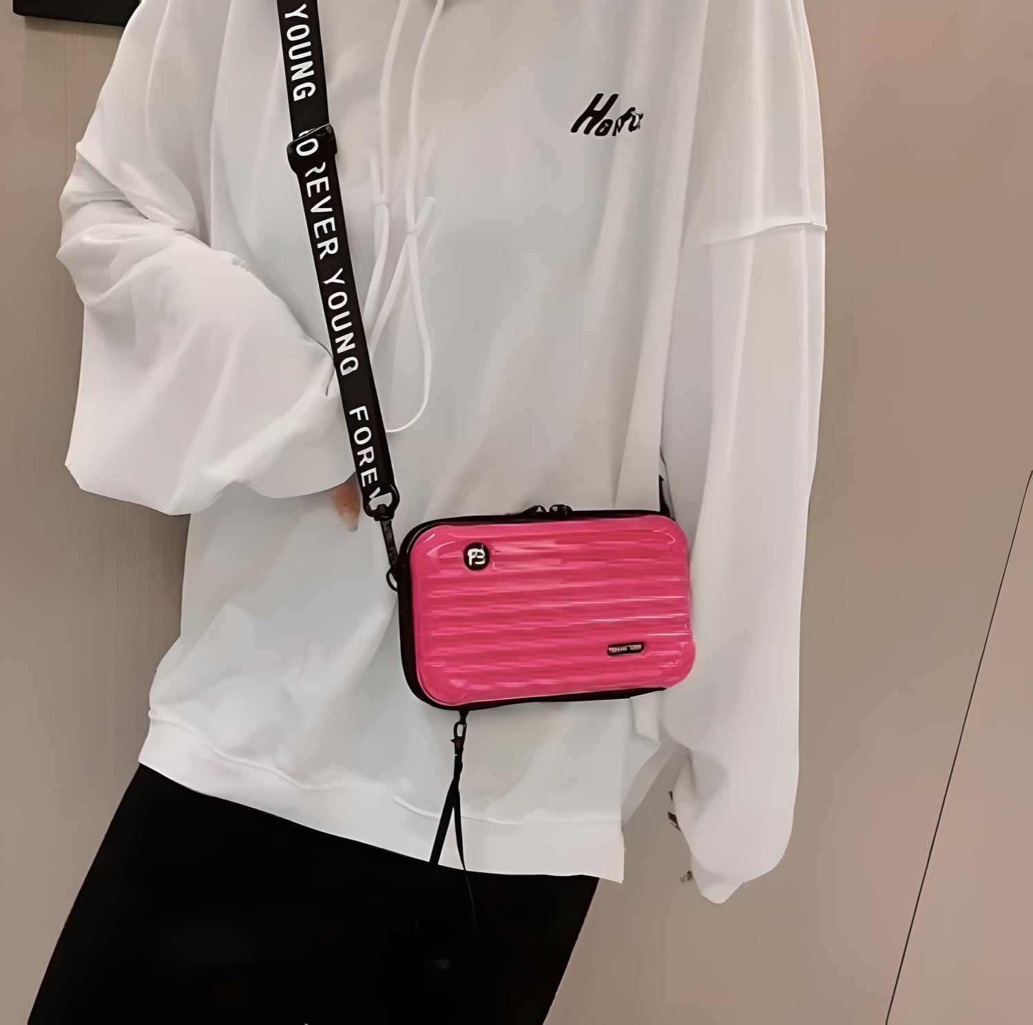 Person wearing a white hoodie and black pants, holding a small pink crossbody bag with a black strap. Fashionable casual outfit, trendy accessories.