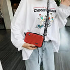 Person wearing a white "Championship" graphic sweatshirt and jeans, holding a red mini suitcase crossbody bag with a "Forever Young" strap.