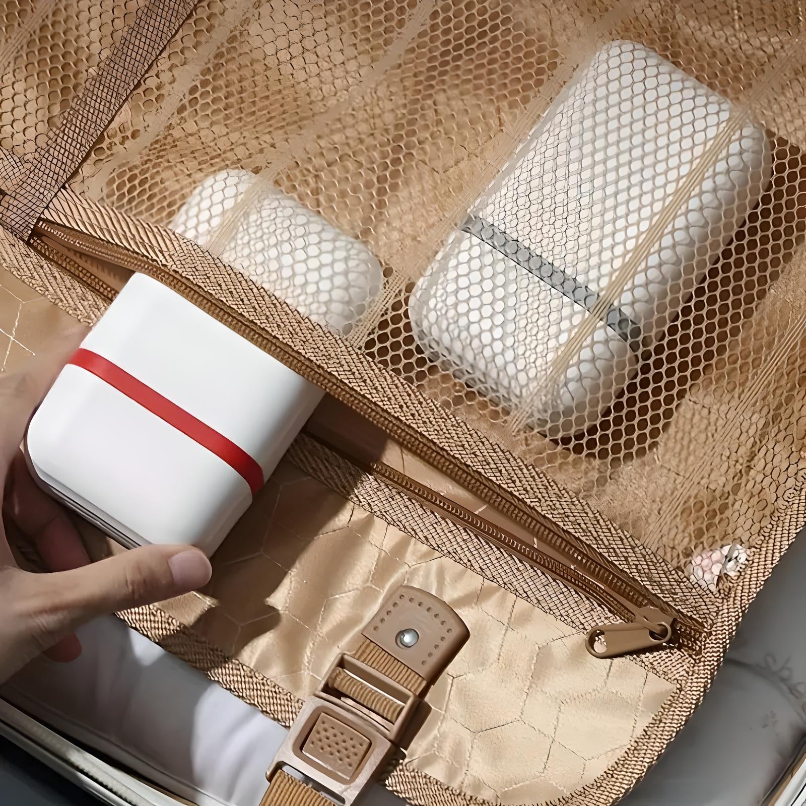 Hand holding a compact white travel organizer with a red stripe, placed in a beige mesh suitcase pocket, alongside other neatly packed items.